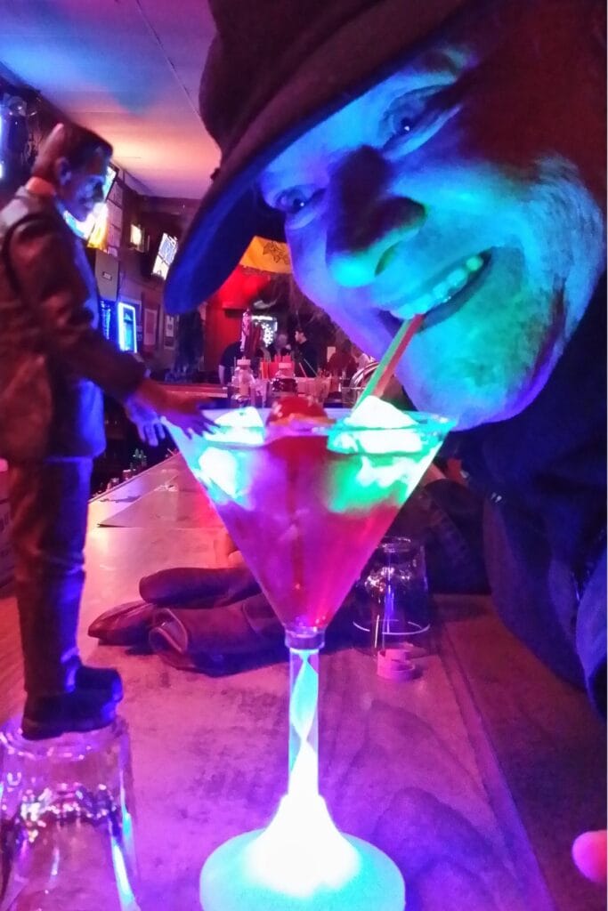 Modern Man in a hat and coat, lit in blue and purple bar light, leaning over a glowing martini glass.