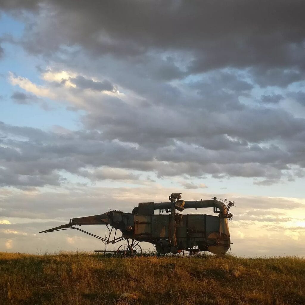 An antique threshing machine standing alone on a prairie hilltop under heavy clouds at sunset.