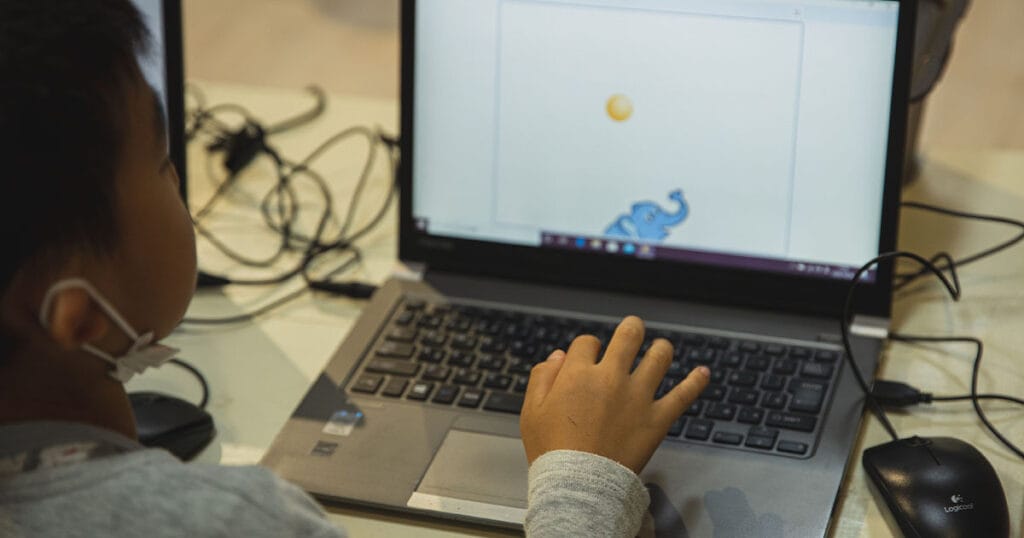 A young child in a face mask working at a laptop in a classroom, watching a simple animation of an elephant and a ball on the screen.