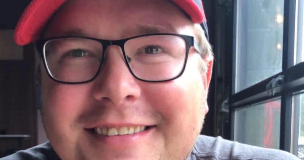 A close-up selfie of a smiling man in a red baseball cap and black-framed glasses, seated by a window.