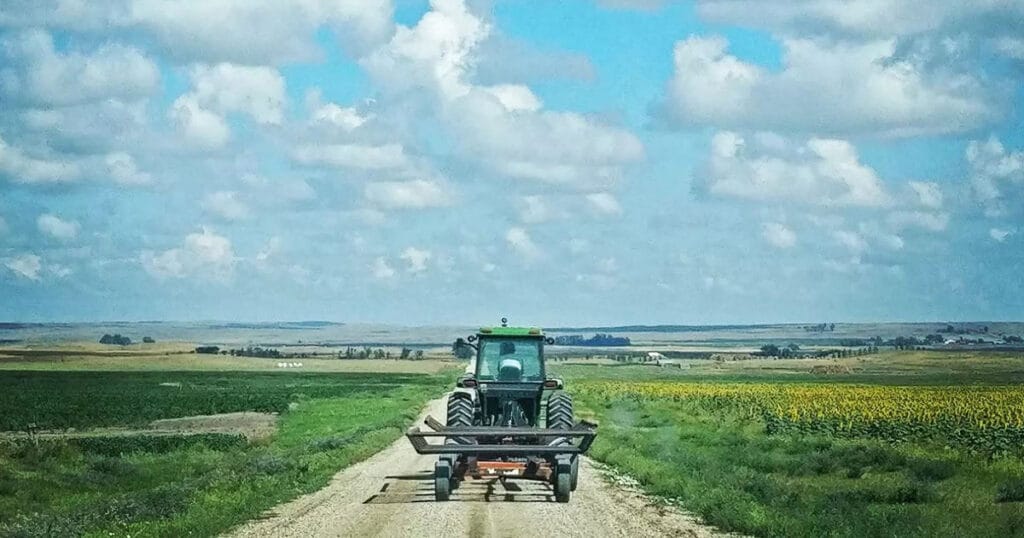 A tractor pulling a hay rake down a gravel road between a green field and a field of sunflowers, under a wide prairie sky.