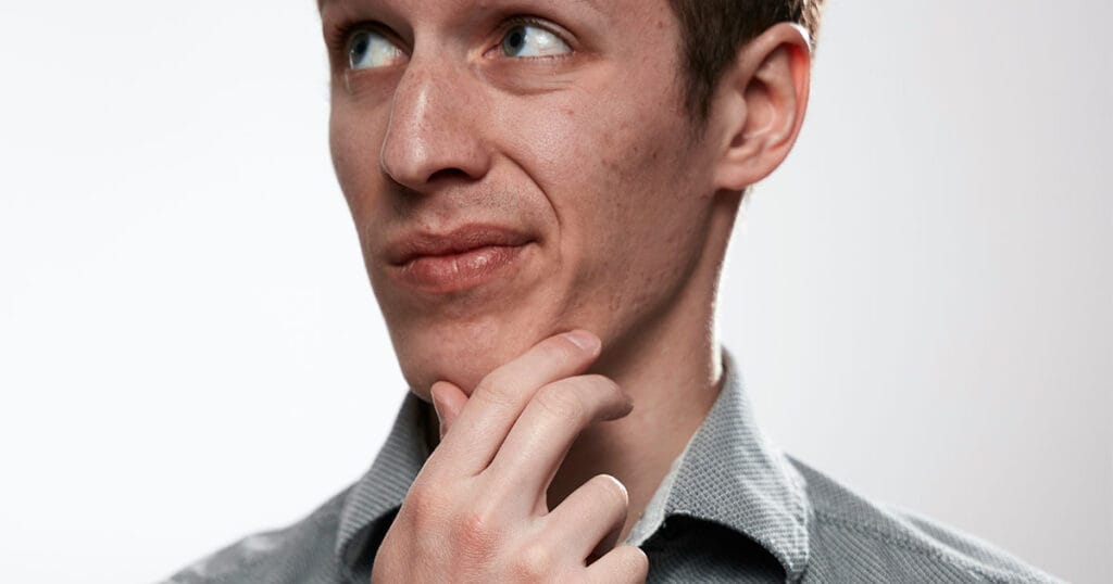 A young man in a grey shirt against a plain background, hand on his chin, glancing sideways with a quizzical expression.