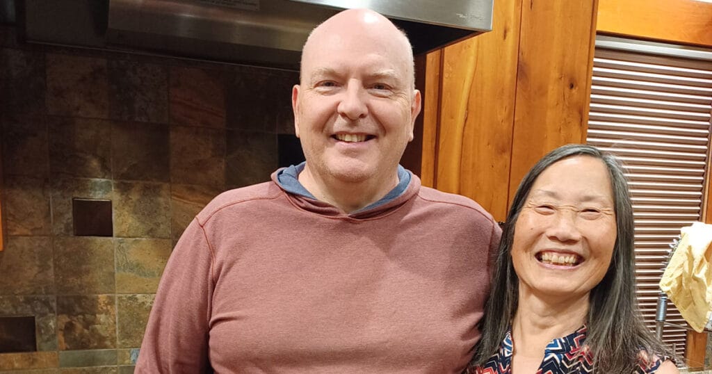 Kevin and Amy Muiderman smiling together in their kitchen, standing in front of a slate tile wall and wooden cabinets.