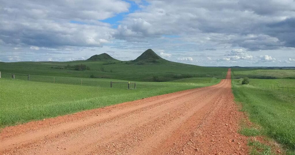 A red dirt road running straight through green prairie toward two rounded buttes under a cloudy sky.