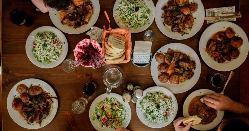An overhead view of a wooden table crowded with shared plates — roasted potatoes, slaws, salads and a basket of bread — as several diners reach in.