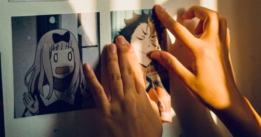Two hands arranging printed anime character images on a wall in warm afternoon light.