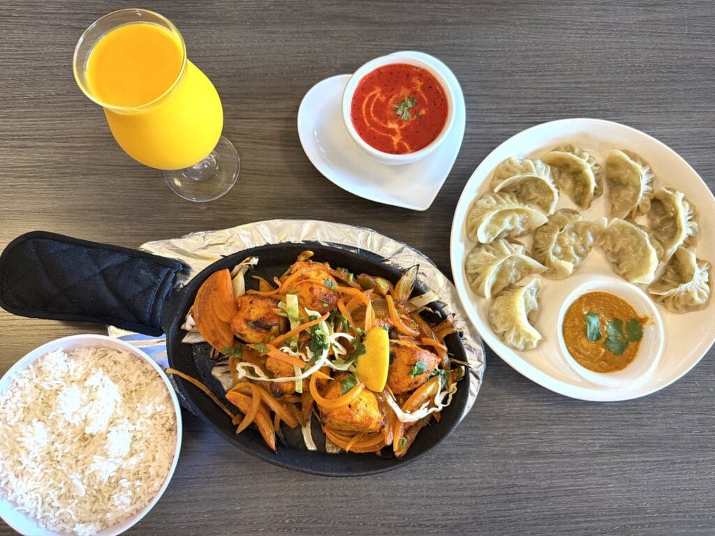 Overhead view of a sizzling platter of tandoori chicken with onions and peppers beside a plate of momos with dipping sauce, a bowl of basmati rice, a cup of red chutney and a glass of orange juice.