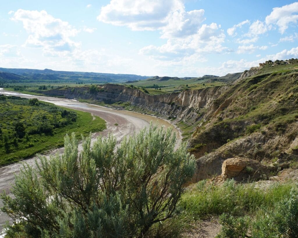 The Little Missouri River curving through the North Dakota badlands, seen from a sagebrush-covered overlook under a partly cloudy sky.