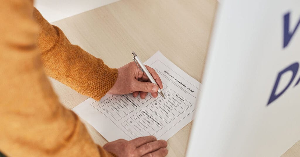 A hand marking an Official General Election Ballot with a pen inside a white voting booth.