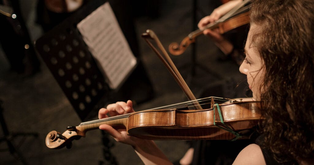A violinist draws her bow across the strings in a darkened concert hall, with a second player and a music stand behind her.